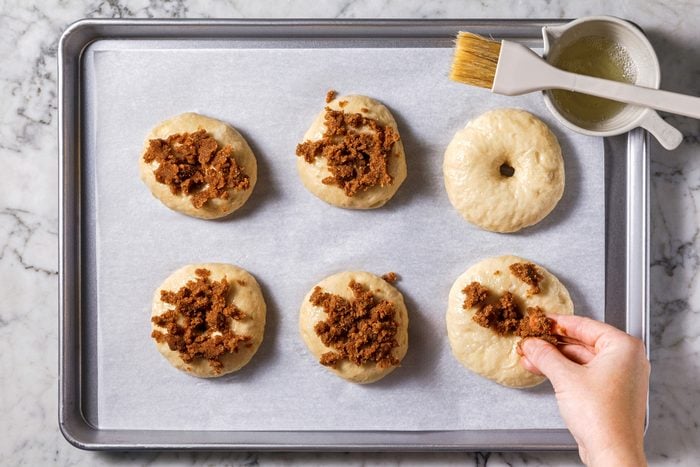 Overhead shot of placing bagels 2 inches apart on greased or parchment-lined baking sheets; brushing with egg white mixture and sprinkling with cinnamon sugar crunch before baking until golden brown, 20-25 minutes, on a marble surface.