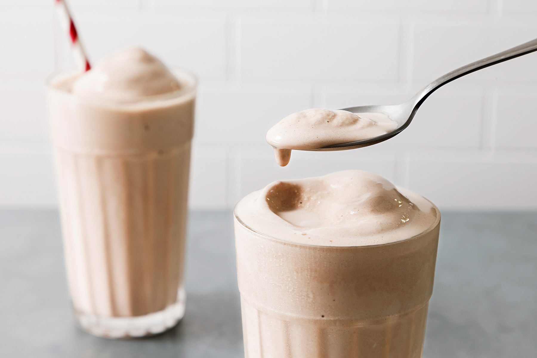 Two tall glasses filled with creamy, frothy milkshakes sit on a gray surface. One shake is paired with a red-striped straw, while a spoon hovers over the other, lifting a portion of the smooth, fluffy mixture. A white tiled wall is in the background.