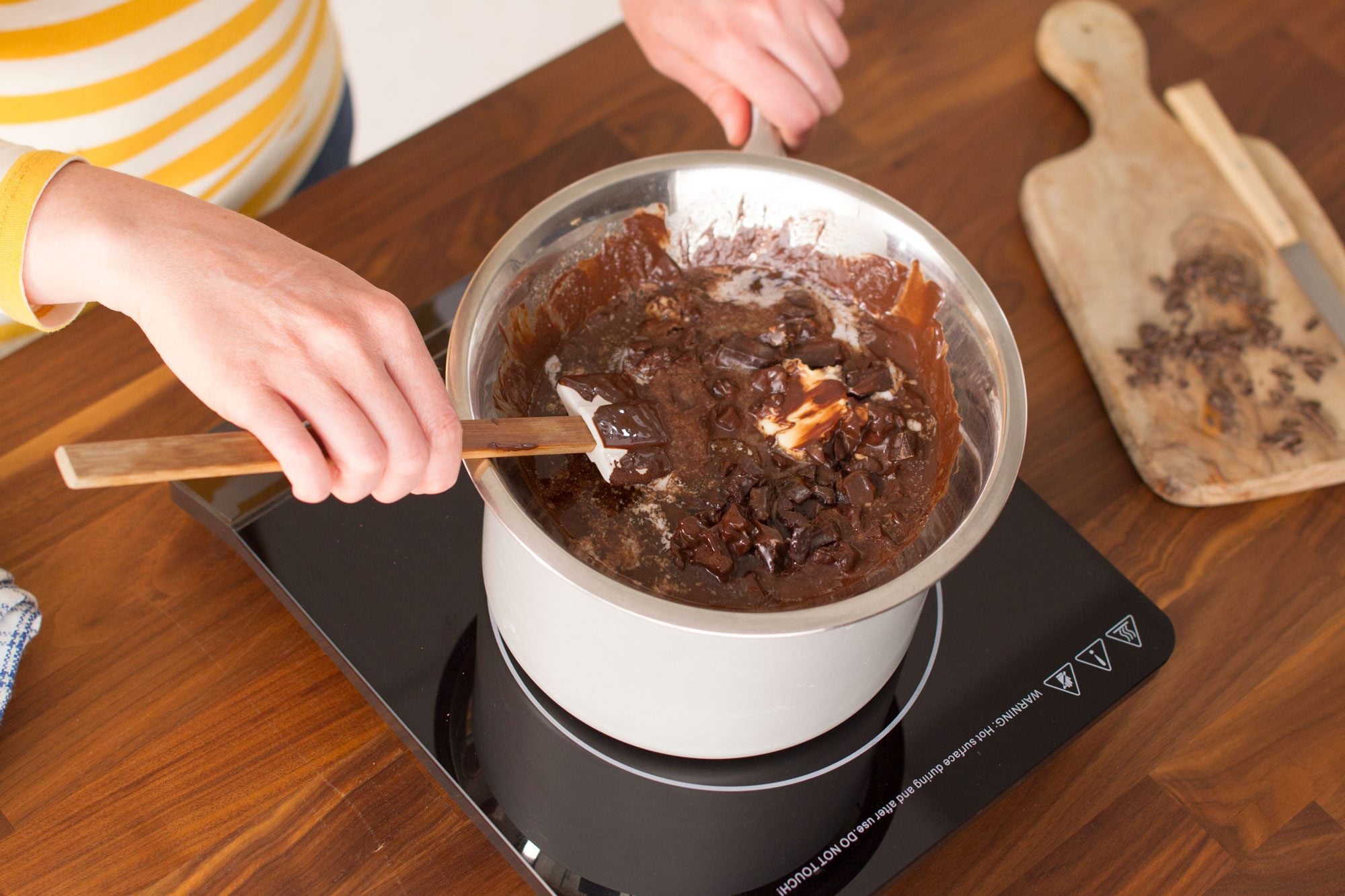 A person stirs a pot of melting chocolate with a wooden spatula on an induction stove. Several ingredients are visible in the mix. A cutting board with chopped chocolate is on the wooden countertop next to the stove.