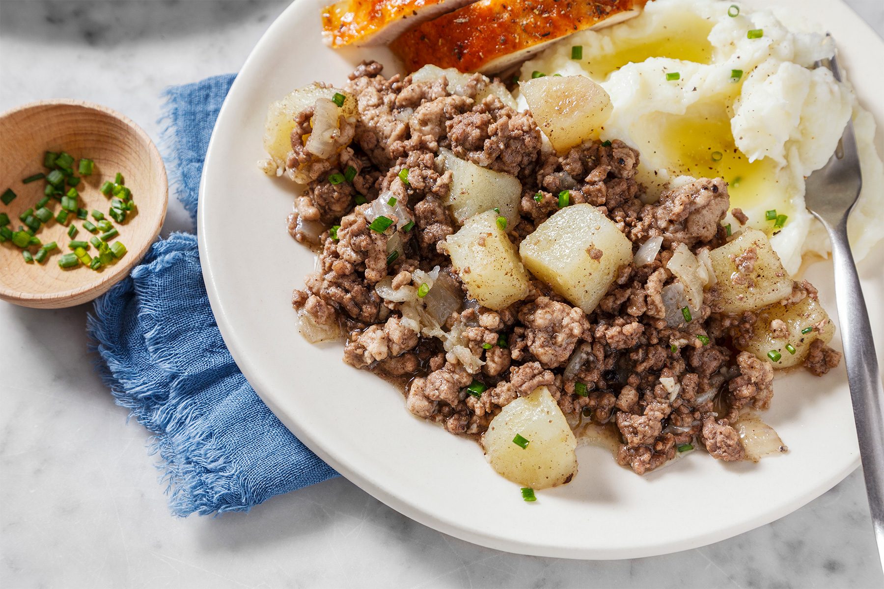A plate of ground beef mixed with cooked onions and potatoes, mashed potatoes with melted butter, and a piece of roasted chicken. A small dish of chopped chives is placed on a blue cloth nearby.
