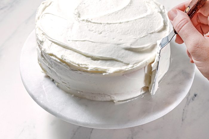 A person uses a spatula to apply smooth white frosting to a round cake on a marble cake stand.
