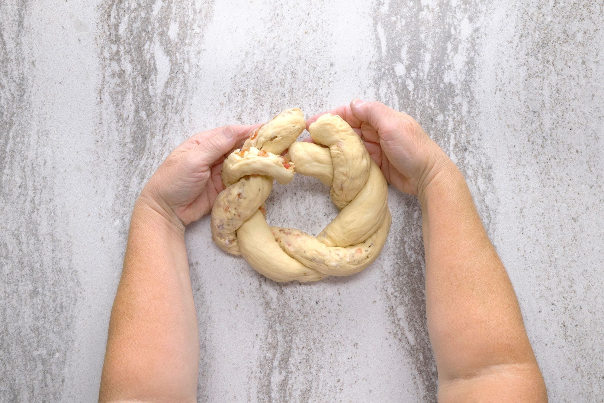 overhead shot of A person is holding a braided dough ring in their hands; the dough is light brown and has small bits of red and yellow inside; the background is grey and textured; the ring of dough is in the center of the image