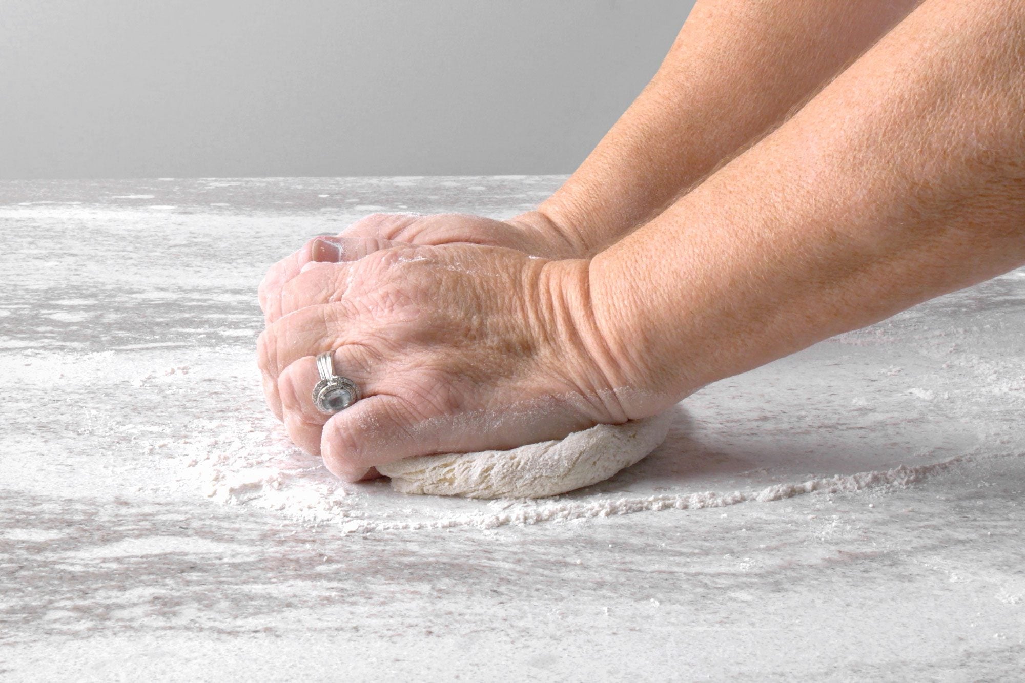 horizontal shot of A pair of hands, one wearing a ring, knead dough on a floured surface; the hands are covered in flour, and the dough is a light brown color; the surface is white and speckled with brown flecks, and there is a trail of flour around the dough