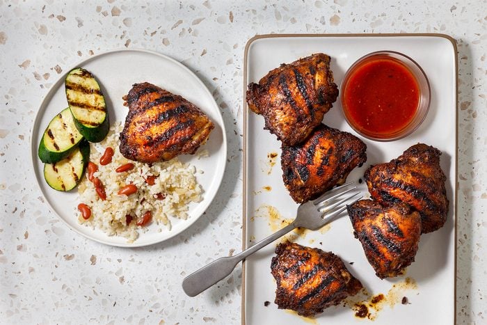 overhead shot of plate of food with grilled chicken, rice, red beans and zucchini, and a plate of grilled chicken with a small bowl of red sauce, a fork, and a few drops of sauce on the plate, all laid out on a white countertop with brown flecks
