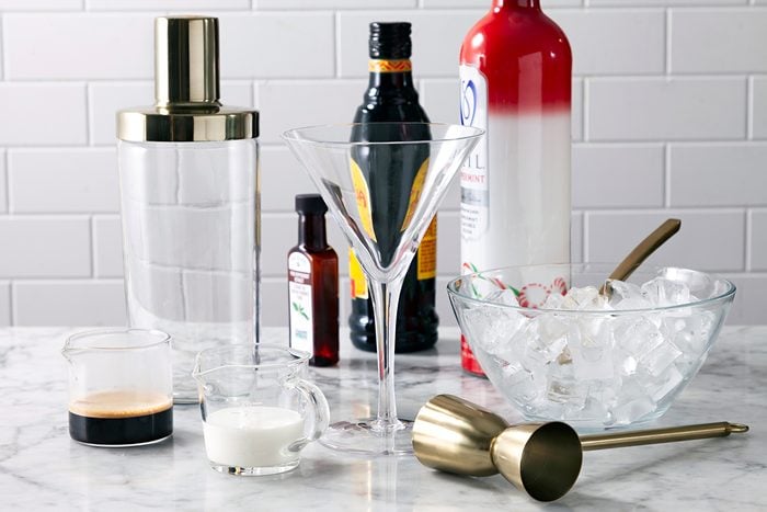 A cocktail-making setup on a marble countertop features a martini glass, shaker, jigger, and a bowl of ice. Bottles of mixers and ingredients like cream and espresso sit nearby, suggesting preparation for a creamy cocktail.
