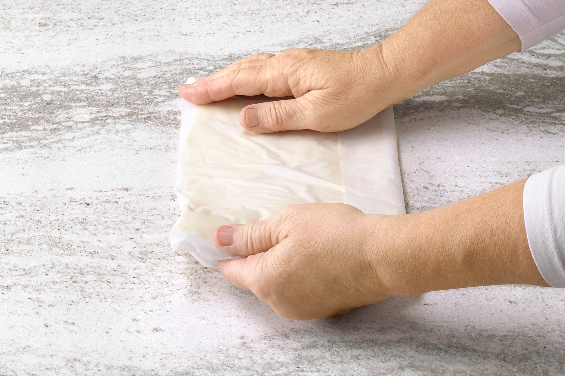 3/4th shot of a person's hands holding a sheet of phyllo dough; the dough is being folded over itself to create layers, and it appears to be translucent and slightly oily