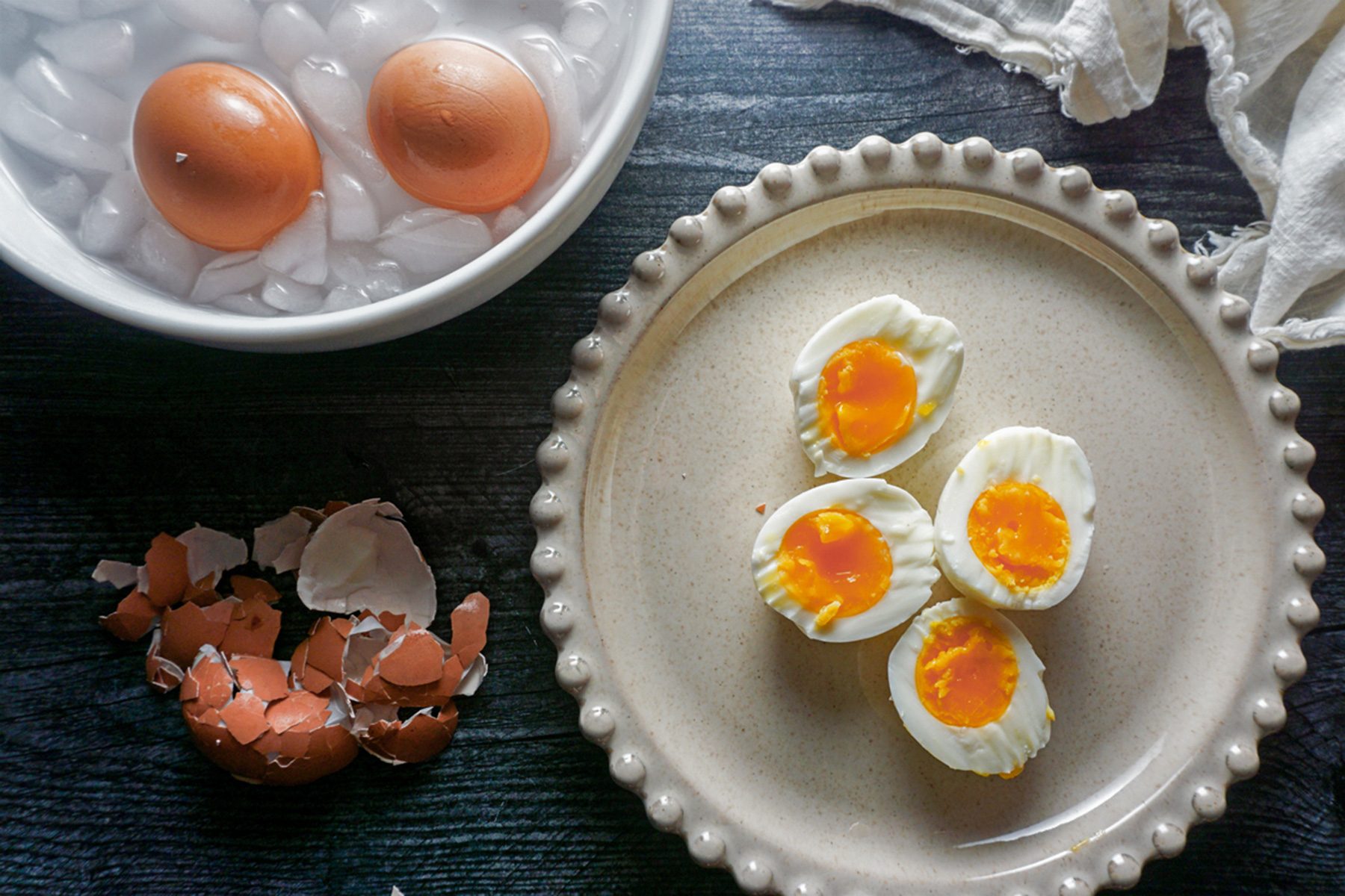 Two soft-boiled eggs are in an ice bath in a white bowl. A plate with four egg halves shows the yolk's soft center. Eggshells are scattered nearby on a dark surface. A cloth napkin is partially visible in the corner.