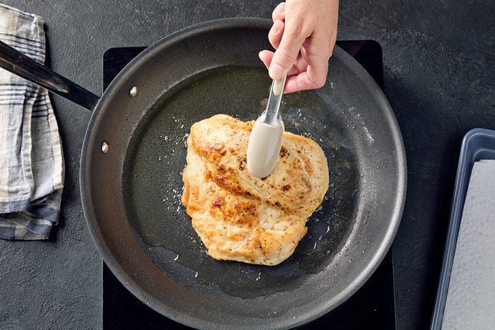A hand uses tongs to flip a cooked chicken breast in a nonstick frying pan. A striped kitchen towel lies beside the stovetop.