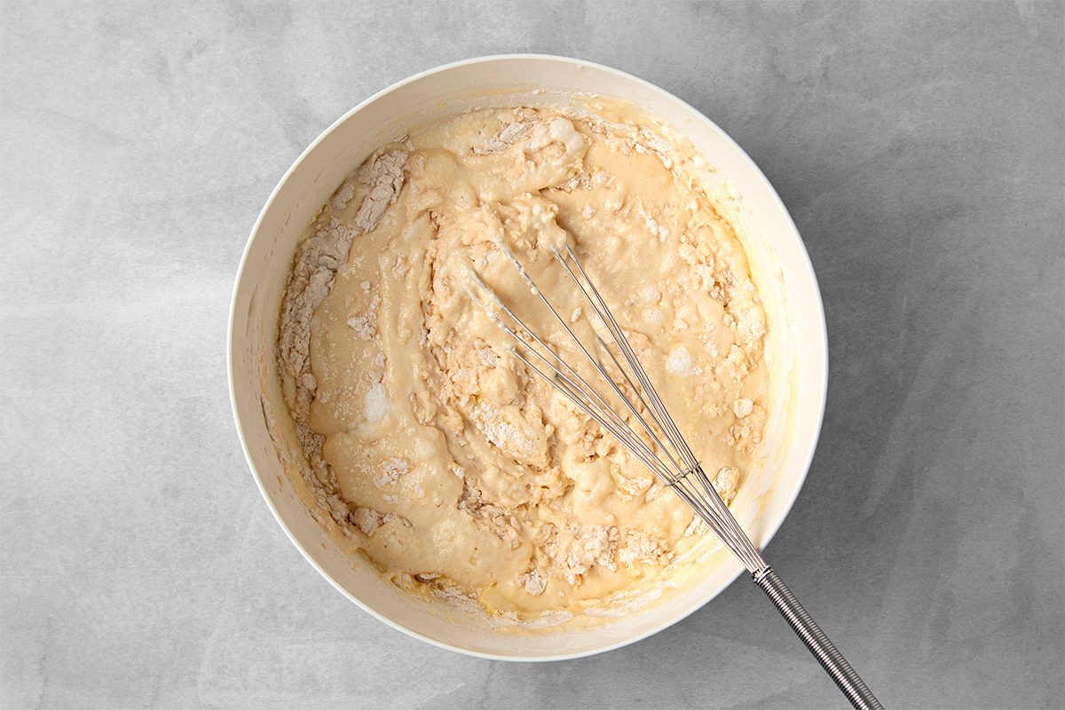 A white mixing bowl containing a thick, off-white batter. A whisk is resting inside the bowl, partially submerged in the mixture. The background is a textured gray surface.