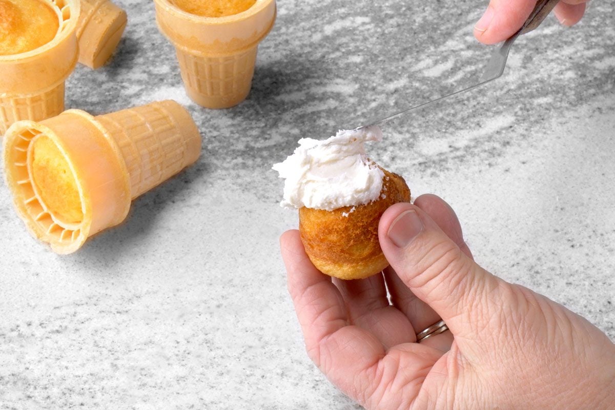 overhead shot of a hand holding a cupcake that has been placed inside of a waffle cone; a knife is used to frost the cupcake with white frosting; three more waffle cones can be seen in the background