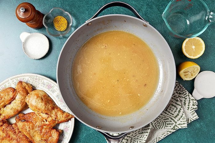 A large pan filled with brown gravy is on a green surface next to a patterned cloth. To the left, fried chicken pieces are on a plate. Surrounding the pan are a pepper mill, small bowls with spices, a lemon half, a glass pitcher, and a salt container.