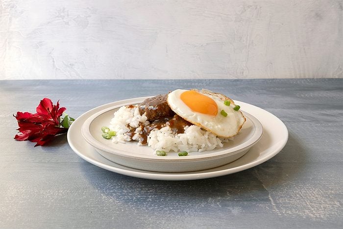 A white plate with rice topped with a brown sauce and a sunny-side-up egg, garnished with green onions. A red flower lies beside the plate on a light blue surface with a textured white background.