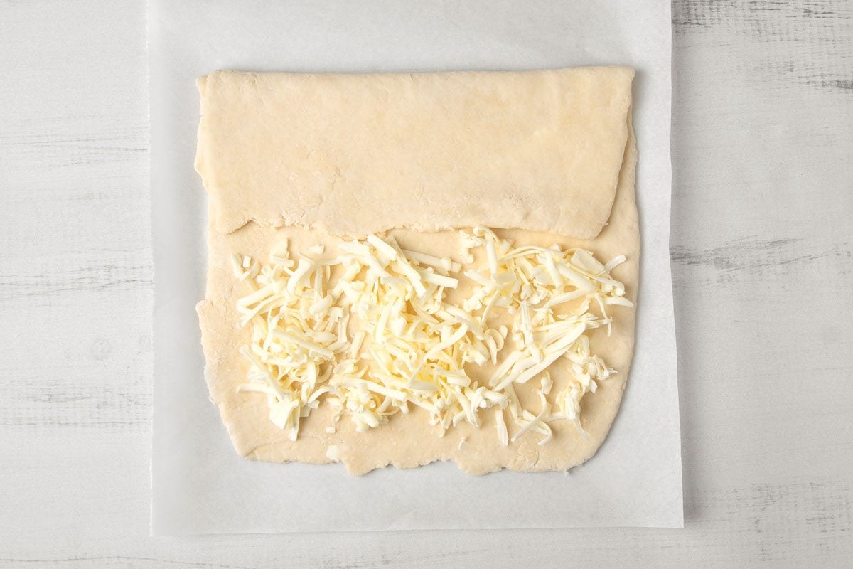 overhead shot of a piece of dough with grated cheese on top of it, on a white wooden surface; it is placed on a piece of parchment paper;