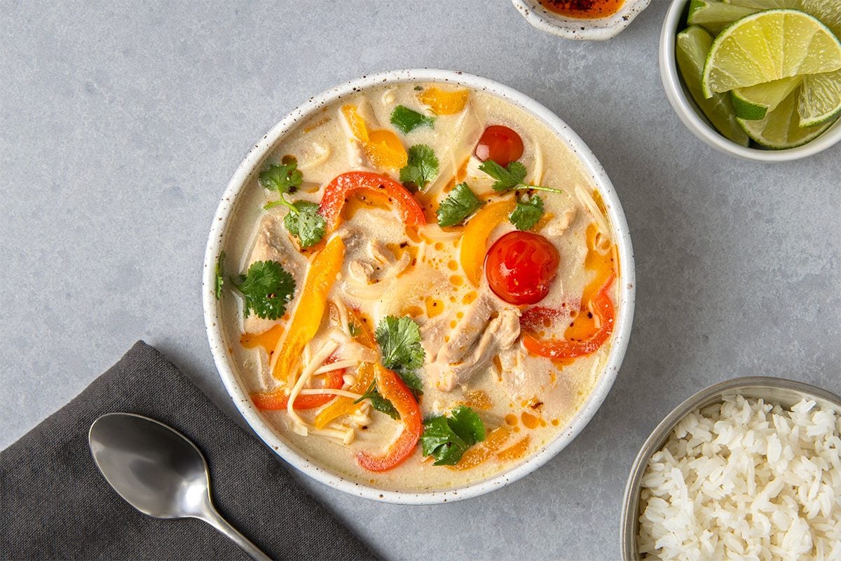 A bowl of creamy soup with chicken, red and yellow bell peppers, cherry tomatoes, and cilantro. Accompanied by a side of lime wedges, a small bowl of rice, and a metal spoon on a gray napkin.