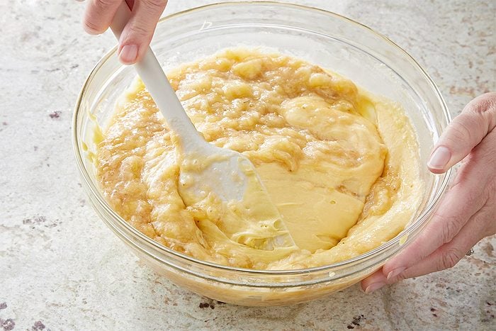 3/4th shot of a clear, round bowl containing a creamy, yellowish a batter, one hand is holding the bowl while another hand is using a spatula to stir the mixture; the background is lightly textured surface