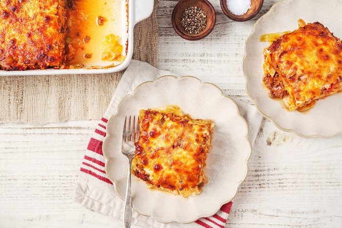 Two plates with slices of lasagna sit on a white wooden table, next to a baking dish containing more lasagna. A fork rests on one plate, and small bowls of salt and pepper are nearby. A red and white striped cloth is beneath the dish.