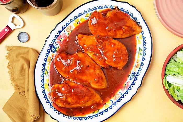 Overhead shot of a dish of Cola Chicken, this particular dish is displayed on a colorful plate with a blue and white pattern and floral accents; there are four chicken breasts covered in a sweet, savory cola sauce; there is a side salad of lettuce in a wooden bowl, and a bottle opener and bottle cap on the table beside the dish; the background is a yellow tablecloth
