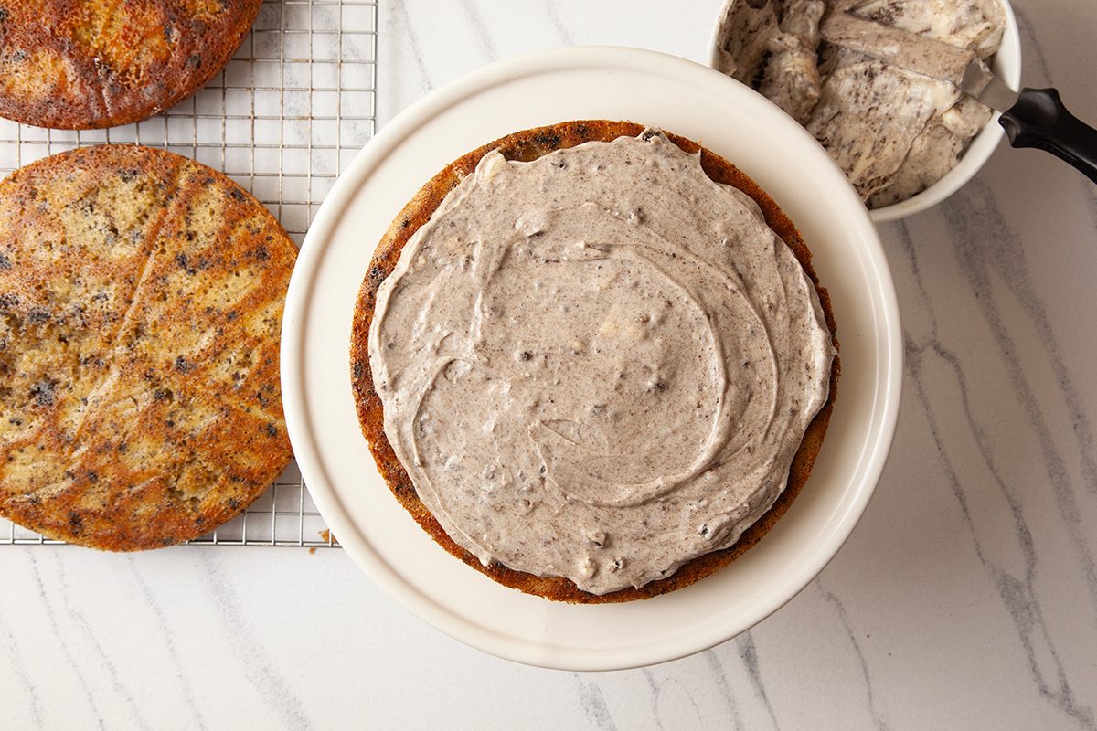 A cake on a white plate is partially frosted with grayish icing. Next to it, a black bowl holds more icing. Another unfrosted cake layer rests on a cooling rack on a white marble surface.