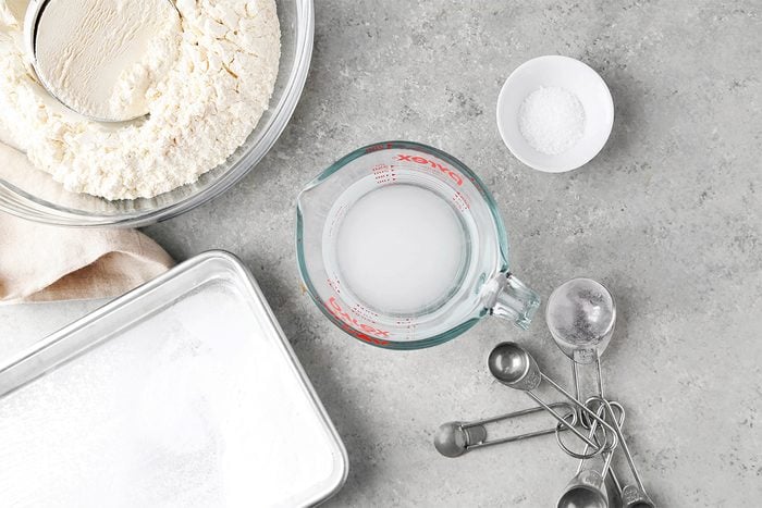 Baking ingredients on a gray surface: a glass measuring cup with milk, a bowl of flour with a sifter, a small bowl of salt, an empty baking tray, and measuring spoons.