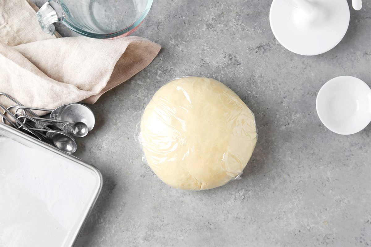 A ball of dough wrapped in plastic sits on a gray countertop. Nearby are a white plate, measuring spoons, a small white bowl, a cloth, and a glass bowl.