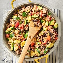 A blue bowl filled with a colorful mix of ground meat, zucchini, tomatoes, black beans, corn, and onions, sitting on a gray surface beside a fork.