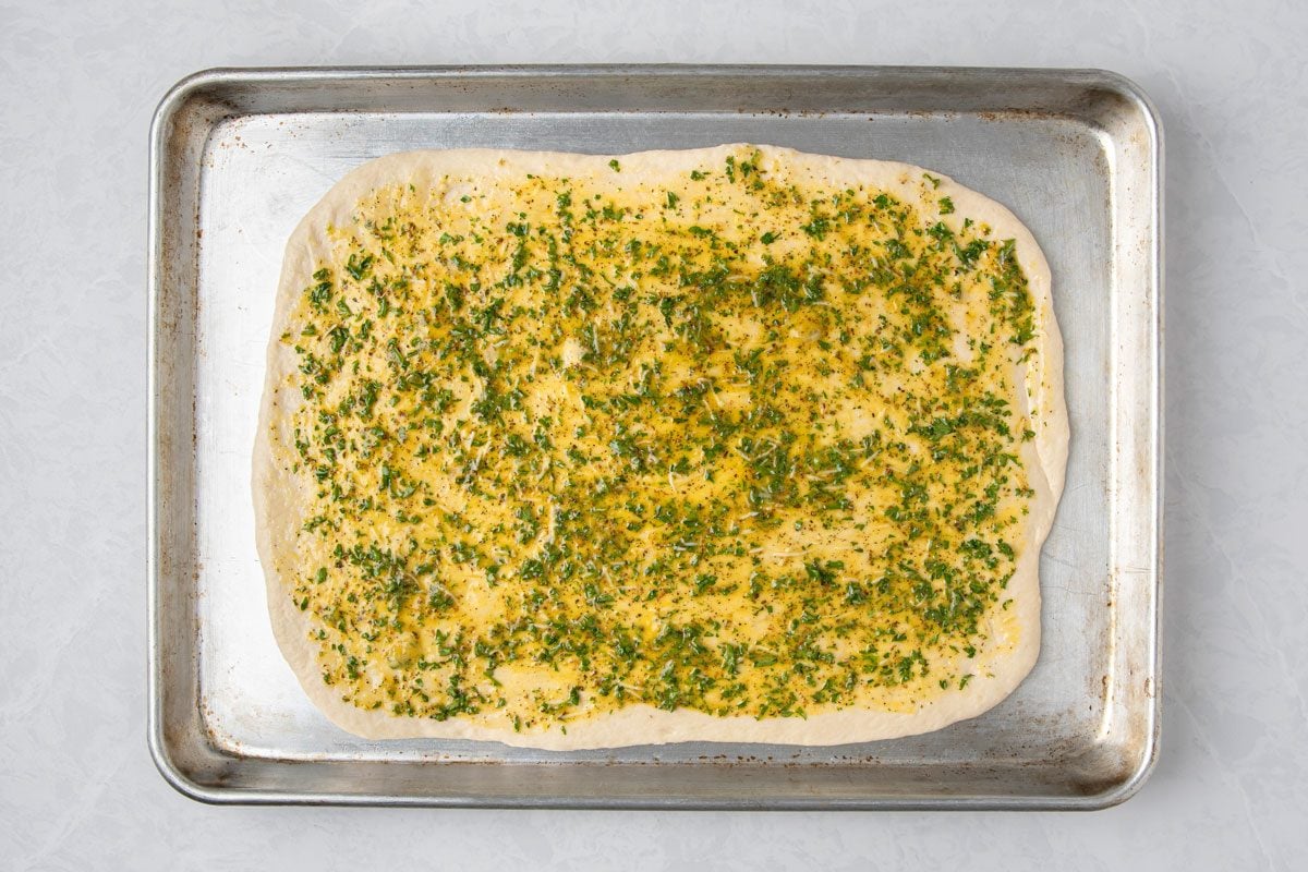 overhead shot of a rectangular piece of dough on a baking tray, the dough is covered with a yellow and green topping that resembles herbs and oil, the baking tray is metallic, a plain white surface background