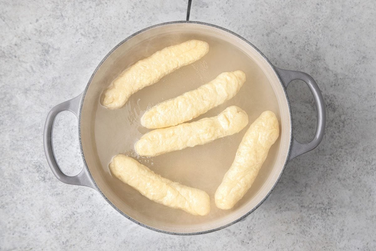overhead shot of several oblong pale yellow pretzel sticks cooking in a pot of simmering water, the pretzel sticks are submerged in the water, the pot is grey with two handles and is placed on a grey countertop surface;