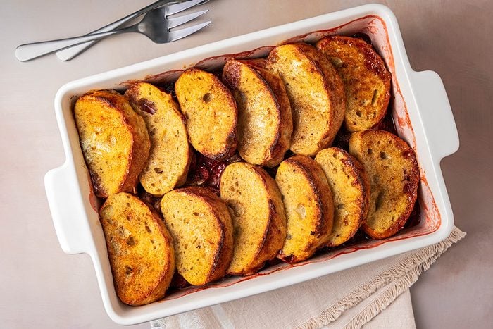 A rectangular baking dish with neatly arranged slices of golden-brown baked bread pudding. The bread slices are slightly tilted, revealing a cinnamon-spiced filling. Two forks rest on the table beside a folded cloth.