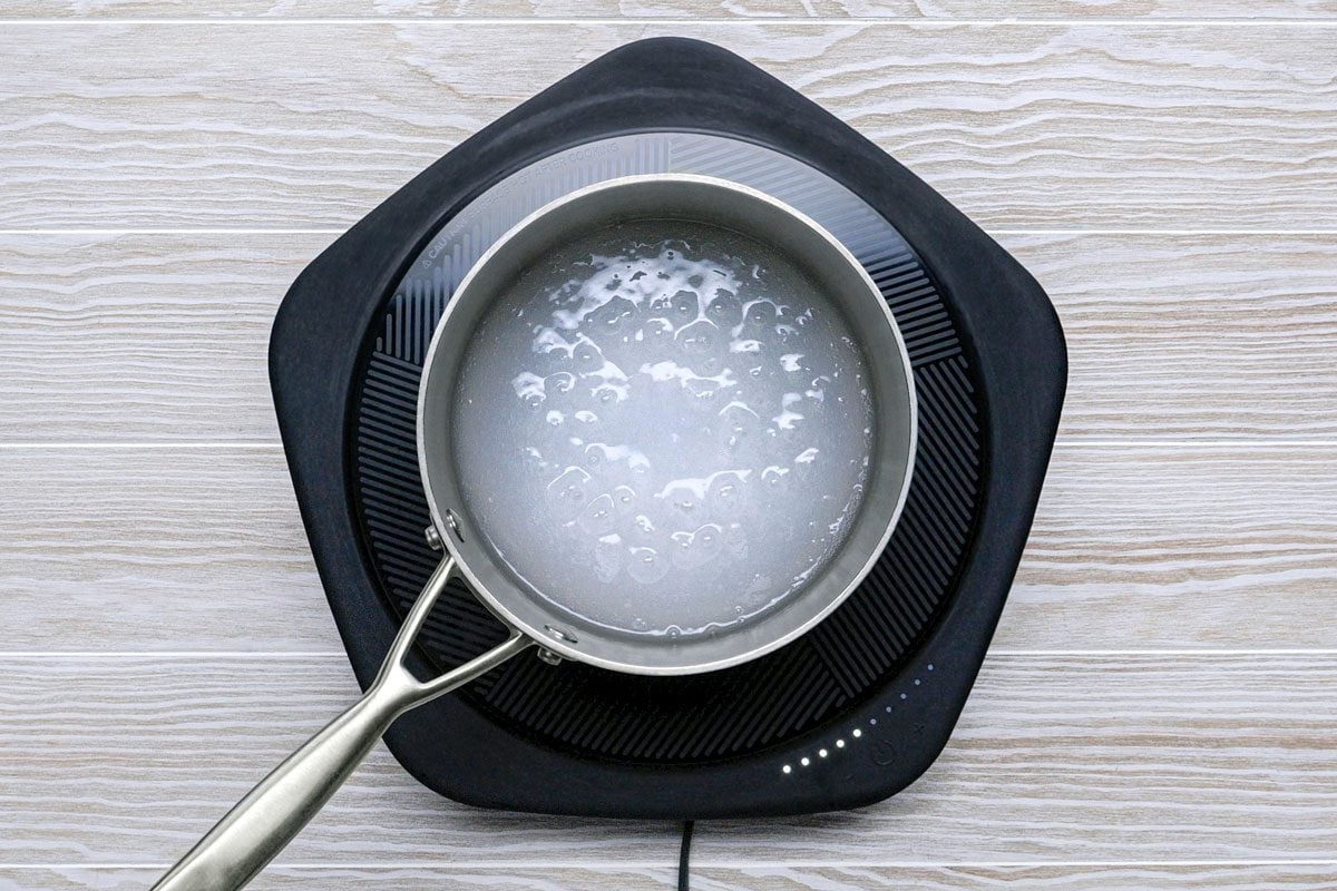 overhead shot of a black electric stovetop with a silver pot sitting on top of it; the pot is filled with water, which is boiling, the stovetop is set on a wooden surface