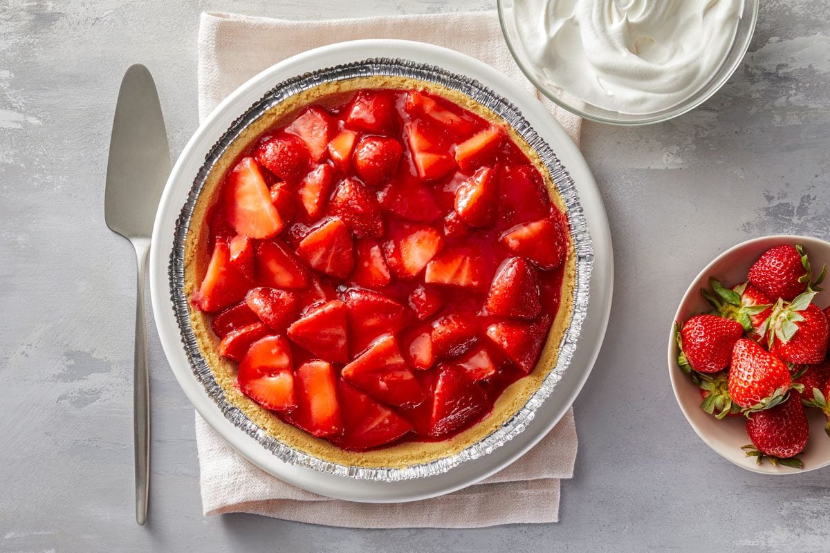 overhead shot of Strawberry Jell O Pie, The pie is positioned on a white plate with a silver-finished crust and is topped with shiny, sliced strawberries soaked in a red gelatinous mixture; Next to the pie, there is a bowl of whipped cream, a small bowl of fresh strawberries, A silver serving knife rests beside the pie