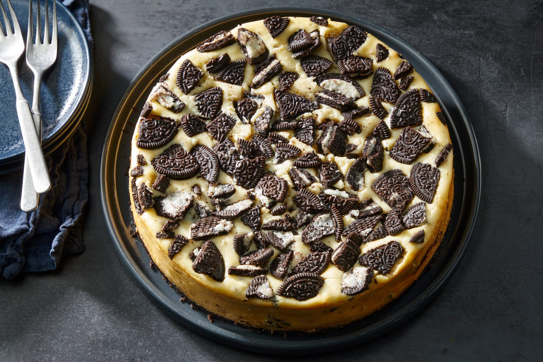 Top view shot of Chocolate Cookie Cheesecake; placed on a large black plate; Empty plates and forks rest on a blue napkin nearby; all set on a dark surface;