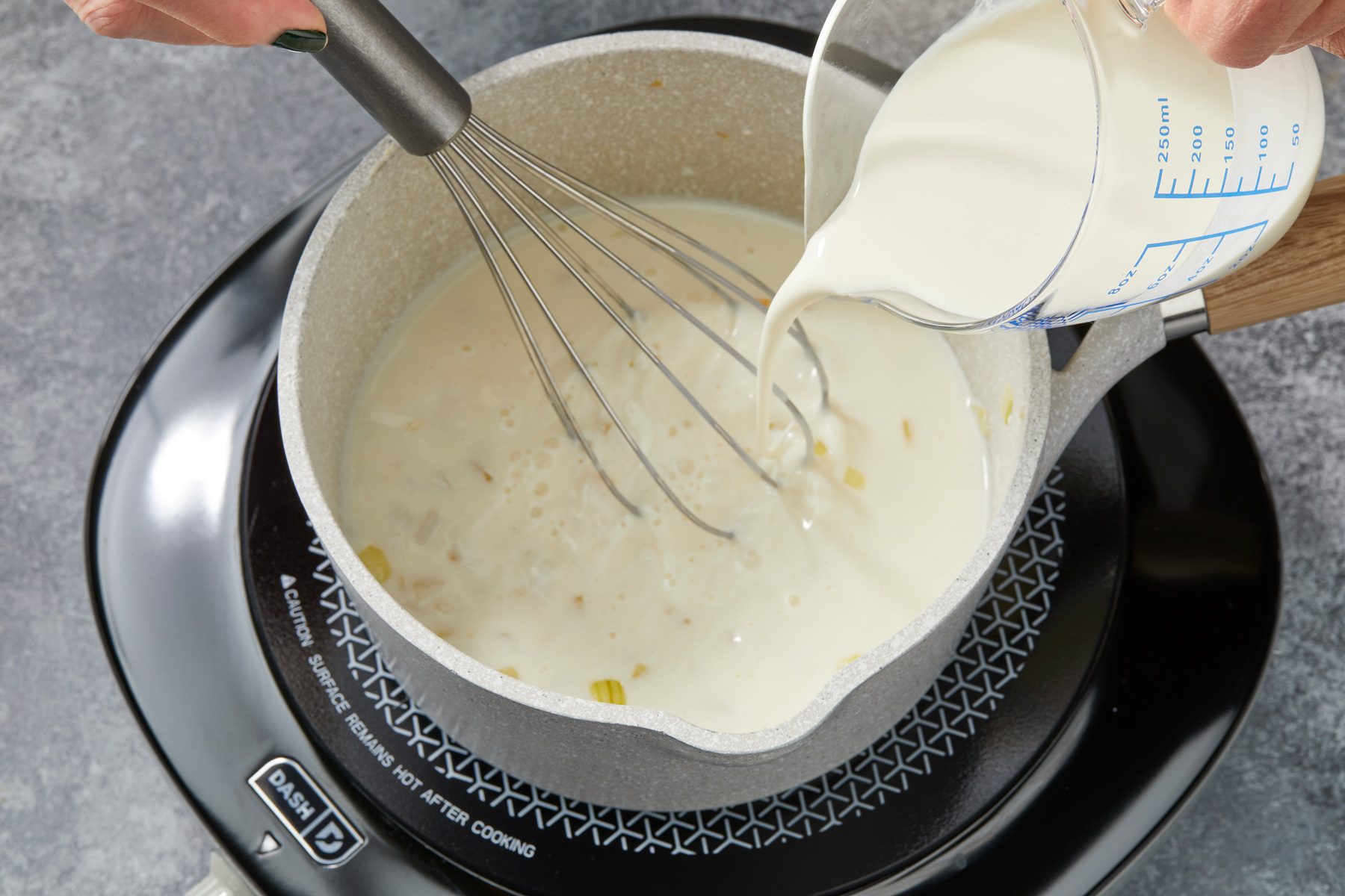 Close shot of the same saucepan; stir in flour until blended; gradually whisk in milk and cream. Bring to a boil; stirring constantly; cook and stir 3-4 minutes or until thickened; induction; all set on a grey surface;