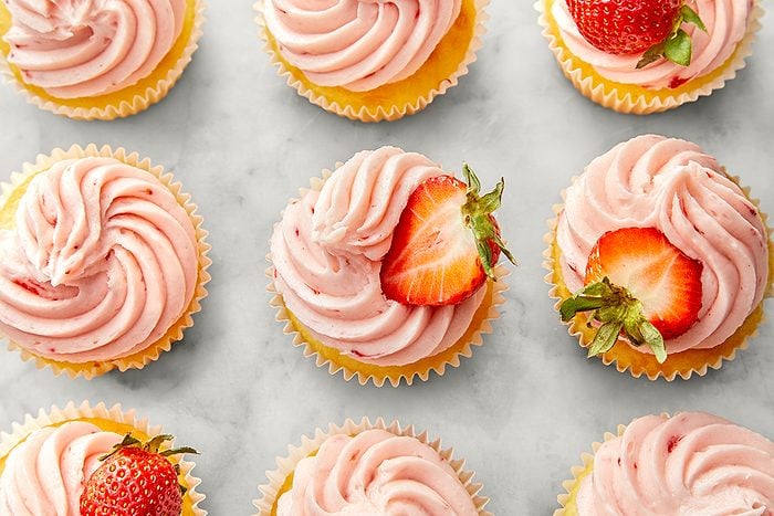 Cupcakes topped with swirls of pink frosting are arranged in rows on a light surface. Each cupcake is garnished with a halved strawberry, adding a touch of freshness to the tempting dessert display.