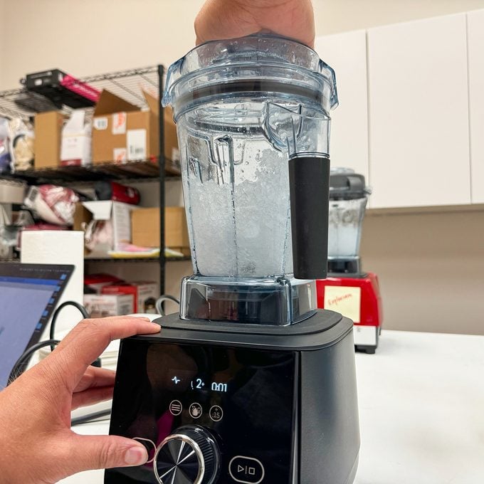 A person demonstrates blending in a transparent blender pitcher, filled with water, on a black base. Shelving and a red appliance are visible in the background.