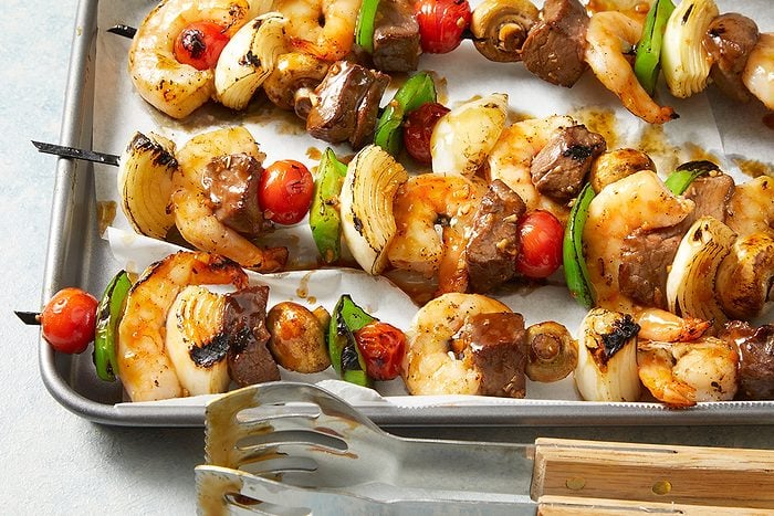 Grilled skewers with shrimp, beef, onions, cherry tomatoes, and green bell peppers on a baking tray, with metal tongs placed nearby.