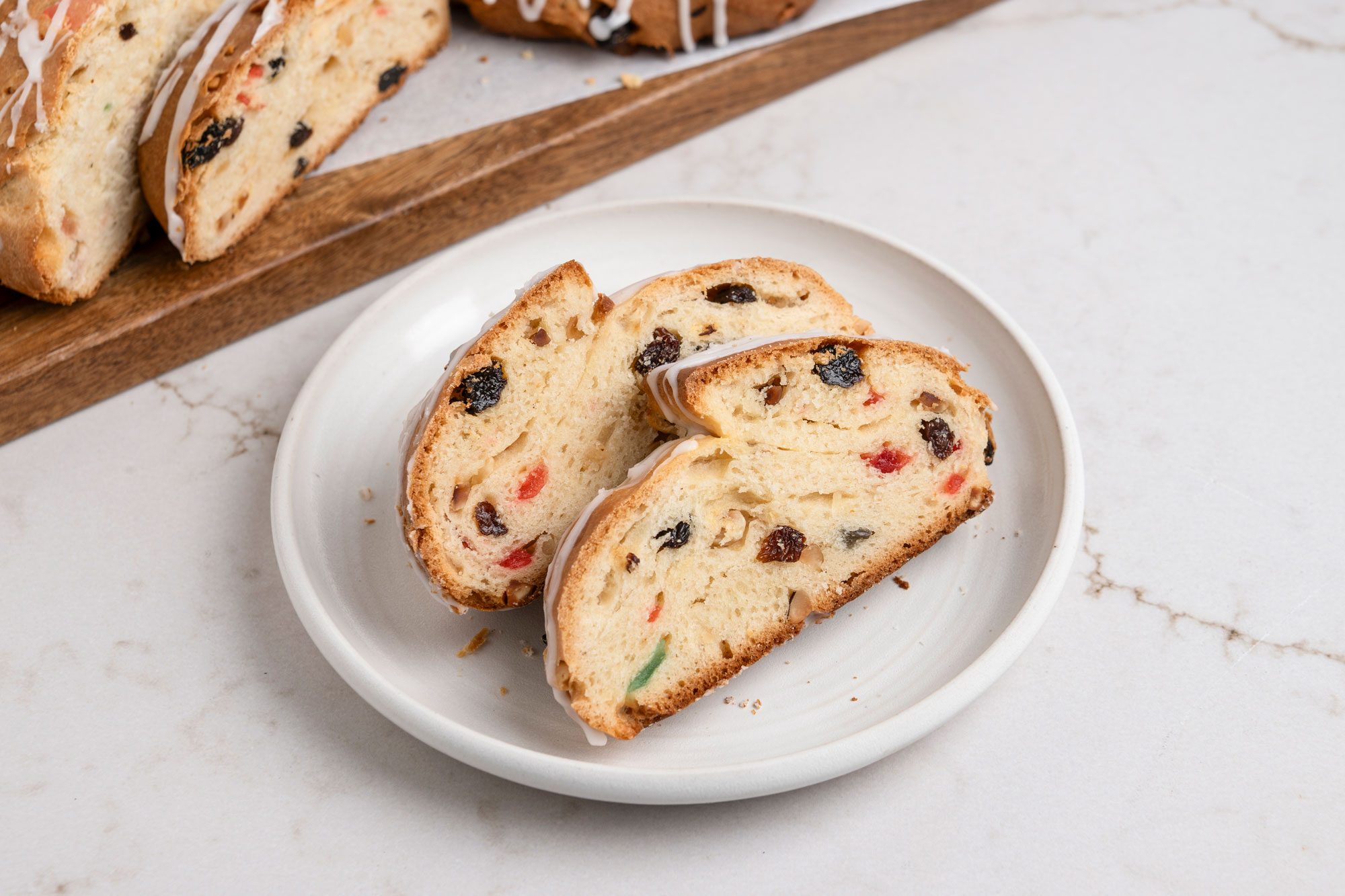 3/4th shot of a Stollen displayed on a simple white plate; A drizzle of white icing decorates the top of the bread, In the background, additional pieces of Stollen can be seen resting on a wooden cutting board