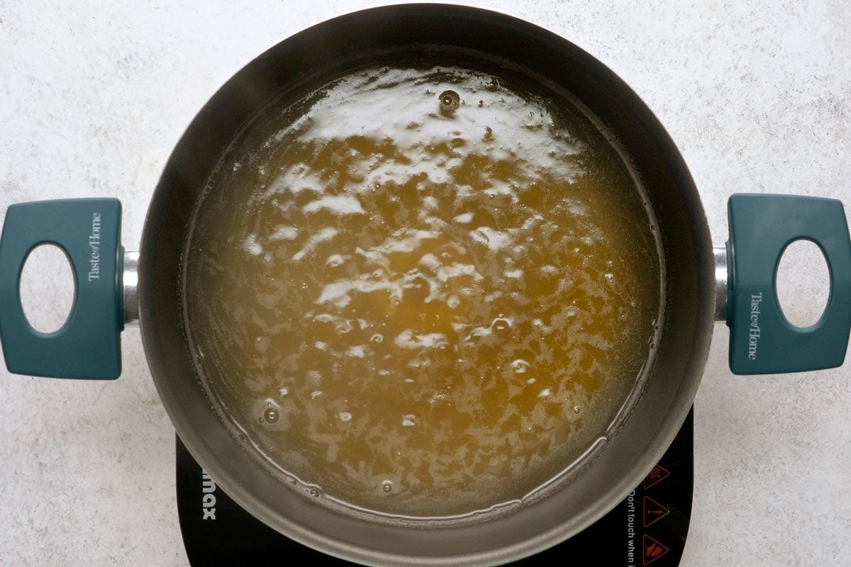 overhead shot of a pot of boiling water is on a stovetop; the water is bubbling and there are air bubbles on the surface of the water; the pot is a stainless steel pot with a blue handle on either side; the background is a light grey counter top;