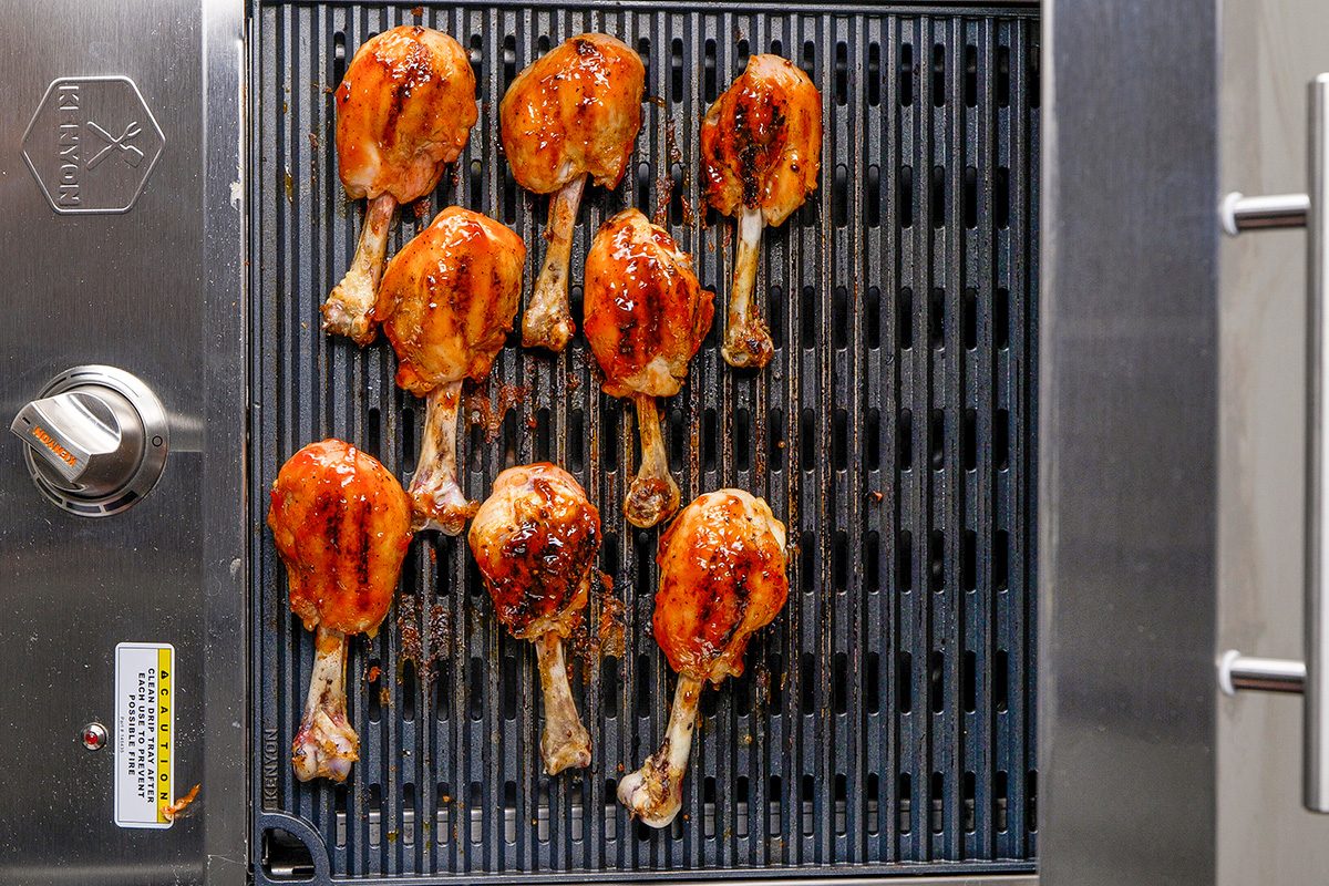 Nine glazed chicken drumsticks cooking on a grill, with visible grill marks and a shiny, caramelized coating on the chicken. The grill is metallic with temperature control on the side.