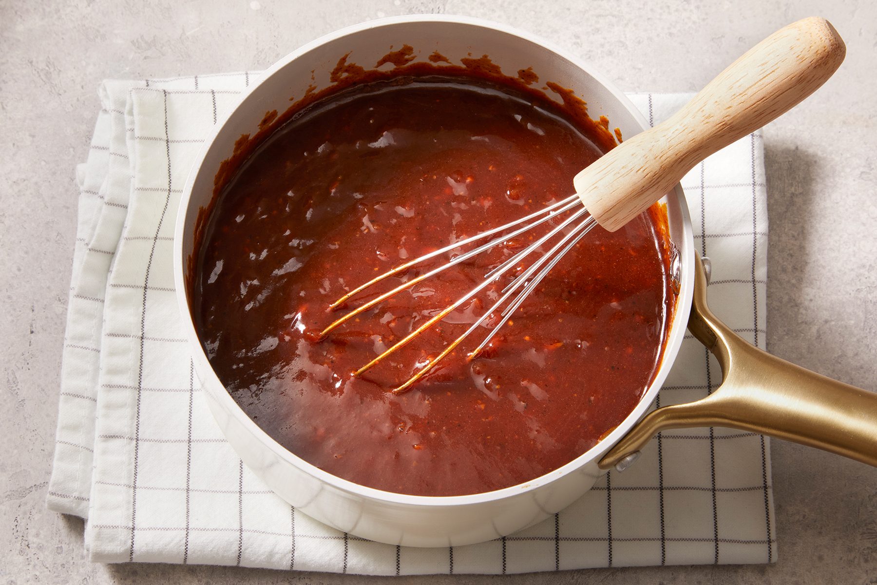 3/4 shot of barbeque sauce kept in a steel pot on top of a check-pattern kitchen napkin on top of a white-grey marble countertop with a wooden whisk inside it.