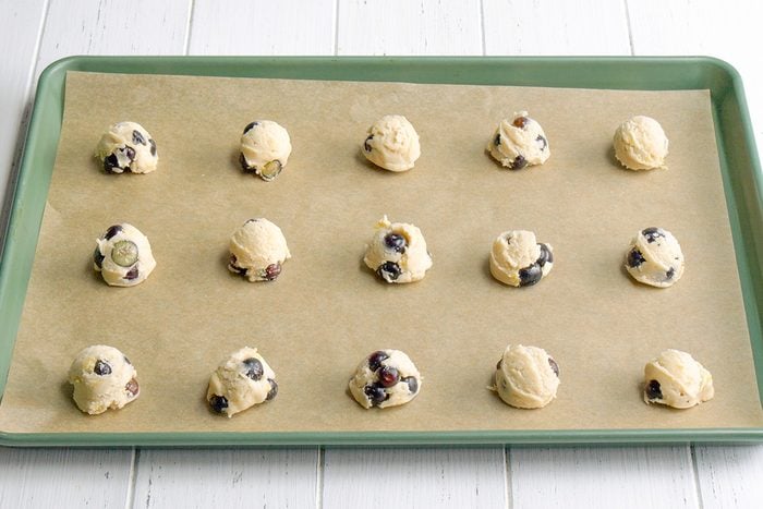 3/4th shot of Fifteen pale, round balls of cookie dough, dotted with blueberries, are arranged on a rectangular baking sheet lined with parchment paper; the baking sheet has a dark green rim and rests on a white wooden surface