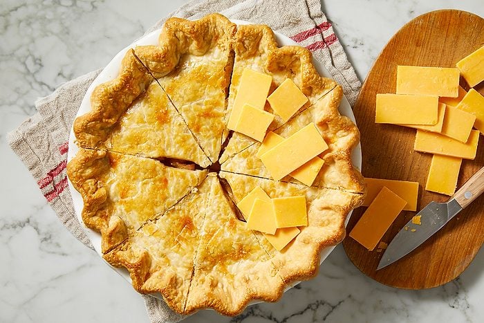 A baked pie with a golden, flaky crust sits on a towel. The pie is sliced, and several slices of cheddar cheese are placed on top and beside it, with a knife and more cheese on a wooden board nearby.