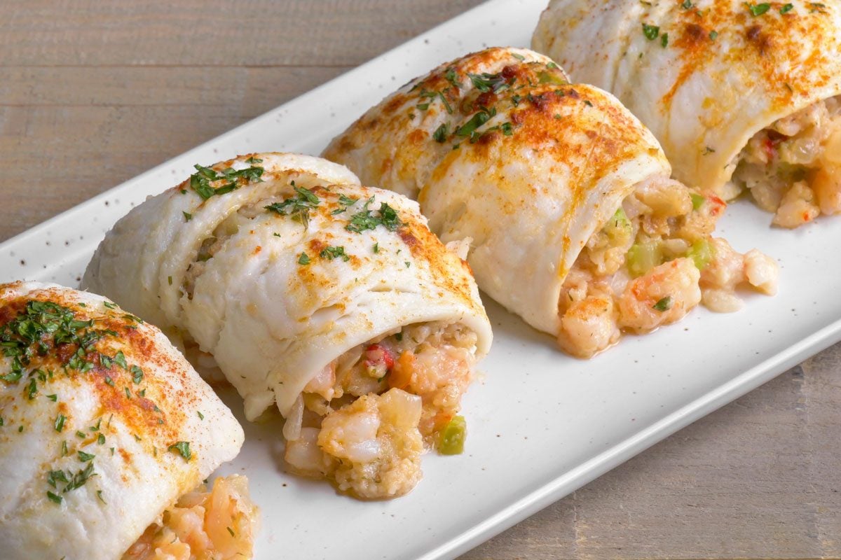 closeup 3/4th shot of Shrimp Stuffed Flounder presented on a rectangular white plate; the dish consists of several pieces of flounder fillet that are neatly rolled or folded; the background is a wooden surface