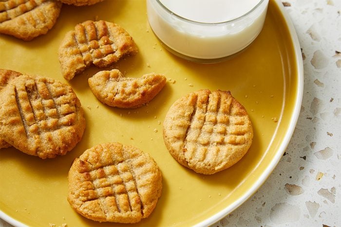 A yellow plate with several peanut butter cookies, some whole and one partially eaten, next to a glass of milk. The cookies have a crisscross pattern on top. The plate is on a light, textured surface.