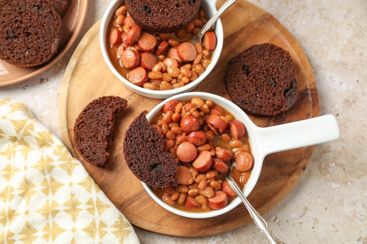 High angle shot of Beanie Weenies in two white dishes with Brown Bread; served on a wooden round tray with two spoons; and a napkin nearby; all set on a marble surface;