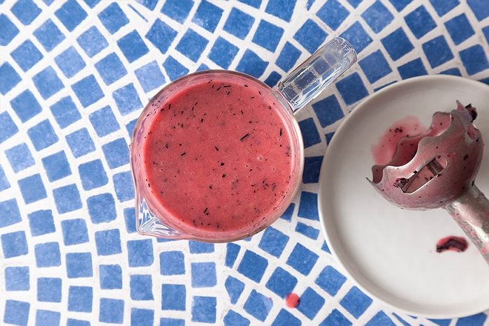 A glass cup filled with a thick, pink berry smoothie sits on a blue and white mosaic table next to a plate holding a partially used ice cream scoop stained with smoothie.