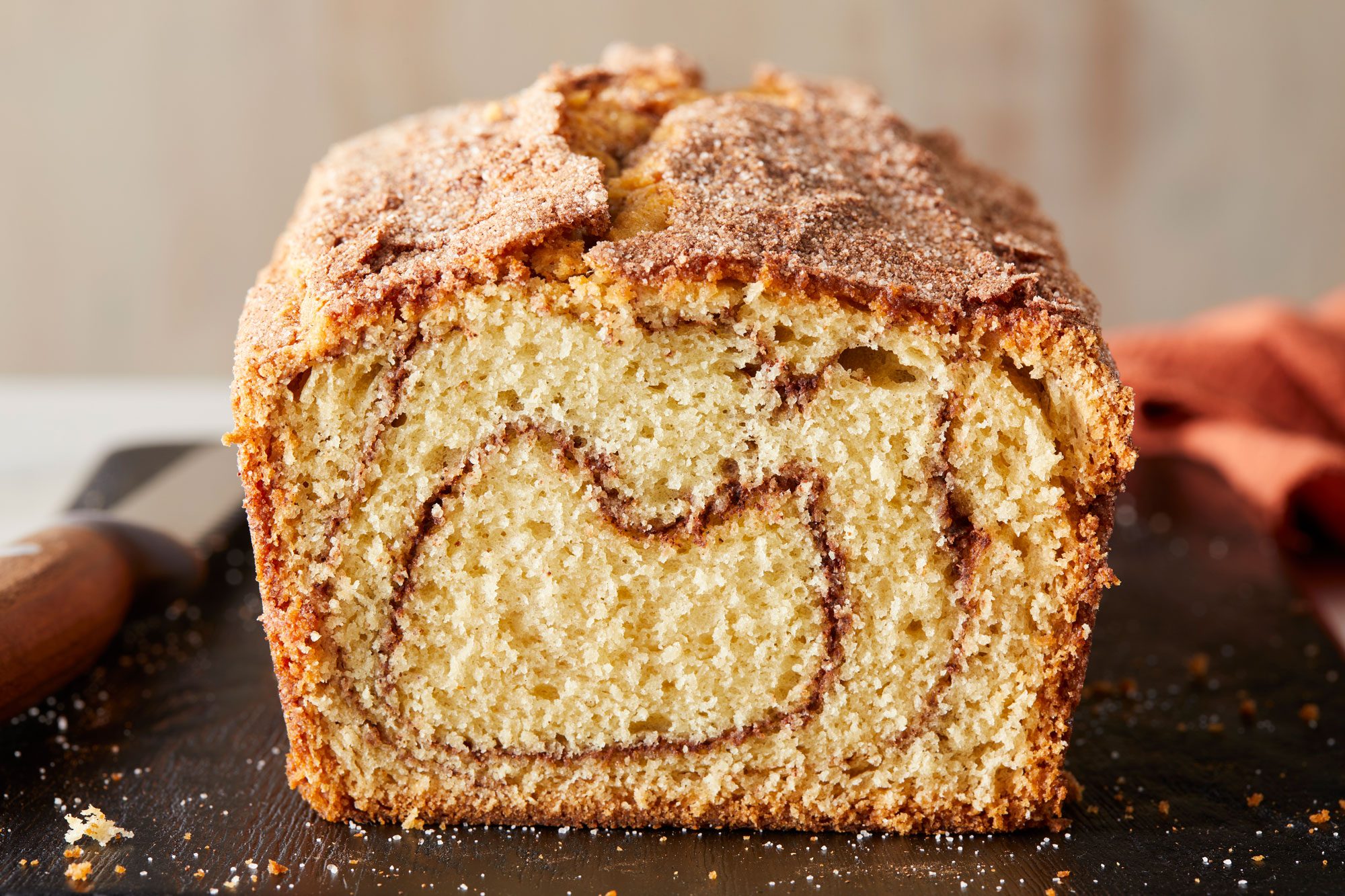 A closeup shot of a sliced loaf of cinnamon swirl bread on a wooden cutting board, showing a spiral pattern inside and a crispy, sugared crust on top