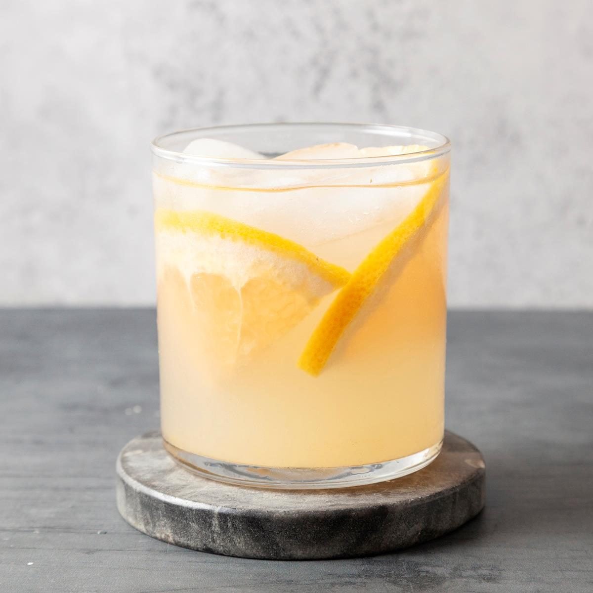 A glass of light yellow citrus drink with ice cubes and lemon slices, placed on a round gray coaster against a light gray background.