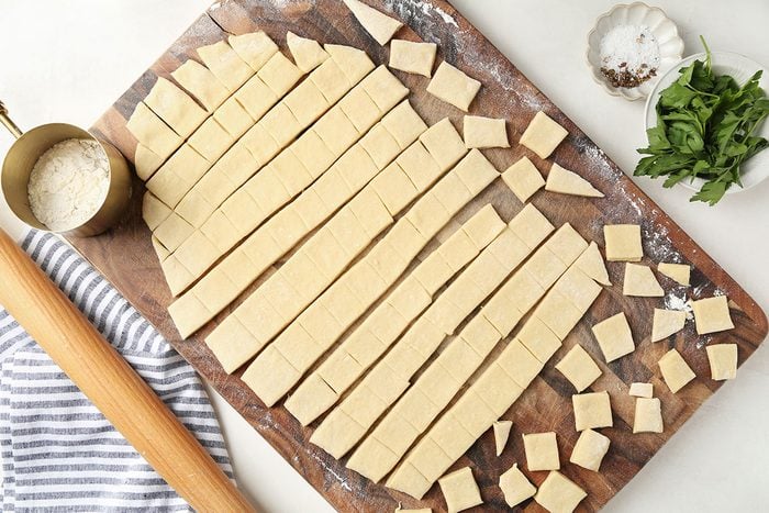 A wooden board with strips and squares of uncooked dough, a rolling pin, a cup of flour, a bowl of salt, and a pile of fresh greens on a white surface.