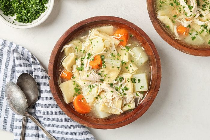 A wooden bowl of chicken noodle soup with shredded chicken, egg noodles, carrots, potatoes, and fresh herbs, beside a striped cloth, two spoons, and a small bowl of chopped parsley.
