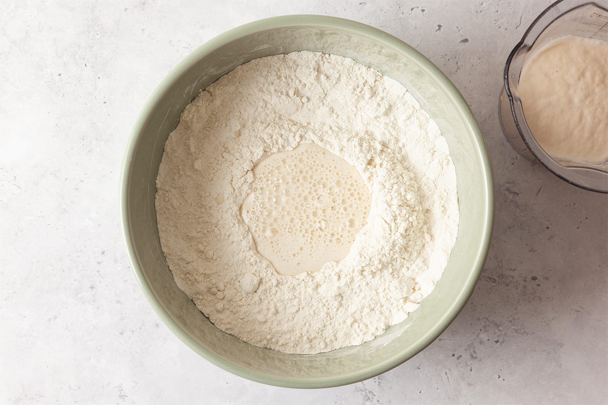A green bowl filled with flour, with a small well in the center containing a bubbling yeast mixture. A second, smaller bowl with another dry ingredient is partly visible to the right.